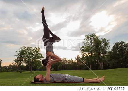 Healthy man lying on grass and balancing woman. Couple doing acrobatic yoga exercise in park Healthy man lying on grass and balancing woman. Couple doing acrobatic yoga exercise in park 93103512