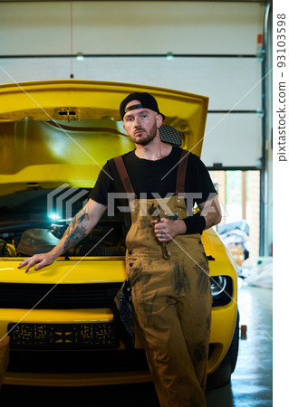 Young serious worker of car maintenance service standing by automobile Young serious worker of car maintenance service standing by automobile 93103598