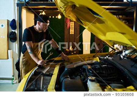 Young repairman with wrench in hand standing by electric car with open hood Young repairman with wrench in hand standing by electric car with open hood 93103608