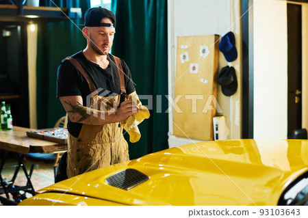 Young repairman in workwear rubbing dirty hands with duster over hood of car Young repairman in workwear rubbing dirty hands with duster over hood of car 93103643