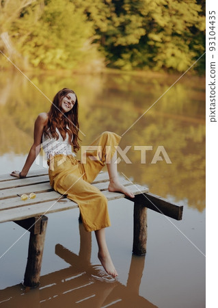 A young happy woman with a hippie smile sits on the lake shore on a bridge wearing eco clothing made of natural materials in harmony with nature in the fall. Travel lifestyle 93104435