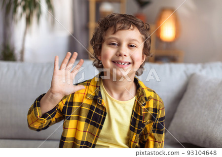 Hello. Camera pov portrait of positive little boy waving hand, video calling or recording blog at home interior 93104648