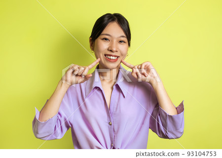 Toothy Asian Female Pointing Fingers At Her Smile, Yellow Background Toothy Asian Female Pointing Fingers At Her Smile, Yellow Background 93104753