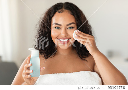 Beautiful black chubby lady cleansing skin with micellar water and cotton pad, making daily beauty routine in bathroom Beautiful black chubby lady cleansing skin with micellar water and cotton pad, making daily beauty routine in bathroom 93104758