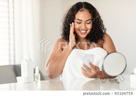 Attractive black bodypositive woman in white towel sitting in bedroom, touching her face and using smartphone Attractive black bodypositive woman in white towel sitting in bedroom, touching her face and using smartphone 93104759