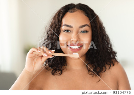 Happy black chubby woman standing in bathroom, brushing her teeth in the morning, looking at camera 93104776