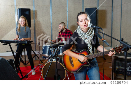 Attractive female soloist playing guitar and singing with her music band in sound studio Attractive female soloist playing guitar and singing with her music band in sound studio 93109794