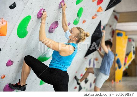 Caucasian woman in face mask exercising on wall in climbing gym during bouldering training. Caucasian woman in face mask exercising on wall in climbing gym during bouldering training. 93109887