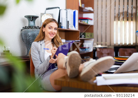 Smiling business woman sitting with feet on table, making notes Smiling business woman sitting with feet on table, making notes 93110214
