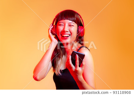 Young asian woman in black tank top wearing red headphones and holding smartphone chatting and listen to music online on the orange background 93118908