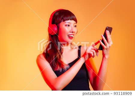 Young asian woman in black tank top wearing red headphones and holding smartphone chatting and shopping online on the orange background 93118909