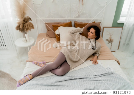 Lifestyle concept. A young middle-aged woman in a sweater lies on the bed and laughs, holds a brown hat in her hands. Lifestyle concept. A young middle-aged woman in a sweater lies on the bed and laughs, holds a brown hat in her hands. 93121973