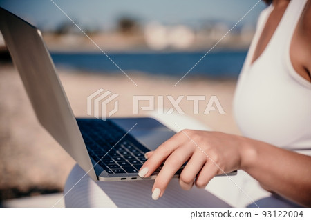 young beautiful woman with dark hair in white overalls sits on the sand near the sea and uses a laptop 93122004