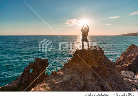 A female tourist, standing with her hands up, reaches the ends of the earth and sees the dragon rock. The girl welcomes the sun. Concept design. A successful woman looks at the sunset with open arms. 93122005