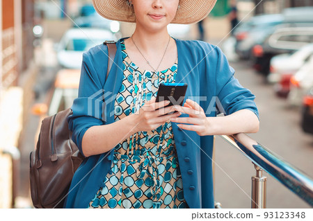 Close up portrait of elegant woman in a hat is typing something on her smartphone. In the background is a street with Parking. Concept of communication and social networks 93123348