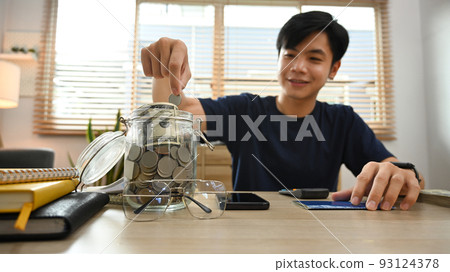 Smiling young asian male putting coin into the jar. Save money for future, retirement and financial planning concept Smiling young asian male putting coin into the jar. Save money for future, retirement and financial planning concept 93124378