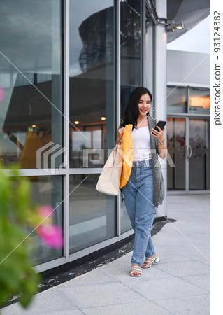 Full length portrait of young woman with shopping bags using smart phone and walking in the city street at the evening Full length portrait of young woman with shopping bags using smart phone and walking in the city street at the evening 93124382