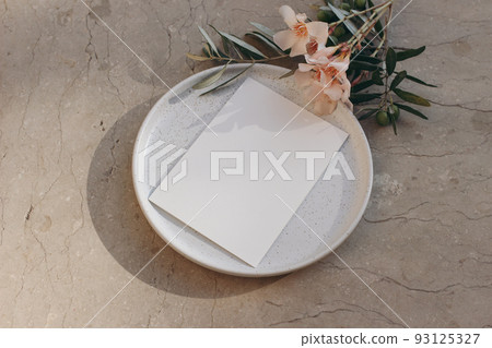 Summer wedding stationery. Mediterranean design. Greeting card, invitation mock up. Speckled white ceramic plate. Apricot oleander flowers, olive tree branches. Beige marble background. Flatlay, top 93125327