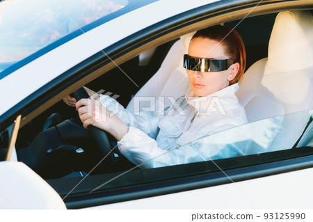Portrait of fashion cyberpunk girl driving a white supercar. Red hair woman with futuristic eyeglasses sits by the car steering wheel. Future technology concept. Selective focus, copy space Portrait of fashion cyberpunk girl driving a white supercar. Red hair woman with futuristic eyeglasses sits by the car steering wheel. Future technology concept. Selective focus, copy space 93125990