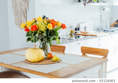 A vase of yellow and orange rose flowers, fresh pumpkin, apple and pear on a kitchen table counter with white modern kitchen background. Autumn home decor for Thanksgiving and Halloween holiday. A vase of yellow and orange rose flowers, fresh pumpkin, apple and pear on a kitchen table counter with white modern kitchen background. Autumn home decor for Thanksgiving and Halloween holiday. 93126063