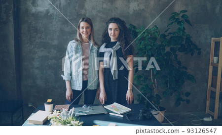 Two female business colleagues are standing together in loft style office, posing and looking at camera. Girls are happy, smiling and laughing 93126547