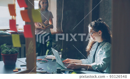 Two attractive girls are working together in modern loft office indoors Curly businesswoman is typing on laptop sitting at table while her colleague is reading book standing near window. 93126548