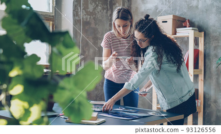 Pretty young women are standing near table, placing colorful pictures on it and shooting flat lay with smartphone. They are sharing ideas about design and smiling. Creative teamwork concept. Pretty young women are standing near table, placing colorful pictures on it and shooting flat lay with smartphone. They are sharing ideas about design and smiling. Creative teamwork concept. 93126556