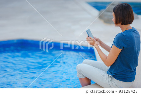 brunette woman using her phone while relaxing by the pool 93128425