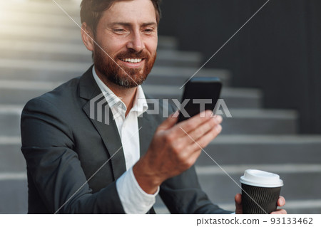 Close up of smiling office worker in suit sitting on stairs with coffee during break and using phone Close up of smiling office worker in suit sitting on stairs with coffee during break and using phone 93133462