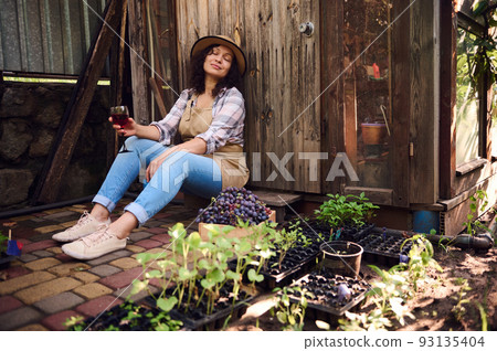 Beautiful peaceful young multiethnic woman wearing a straw hat, sitting at the doorstep of a wooden house in the country house and enjoying a tasting of delicious homemade wine on a warm autumn day. 93135404