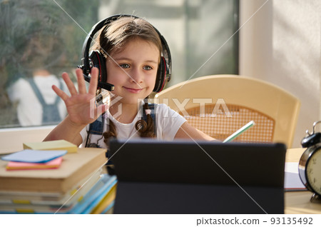 Smart primary school girl wearing wireless headphones, waves with hand her teacher while watching a live online lesson on digital tablet. Education and distance learning for children. 93135492