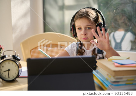 Distance learning. Online education. Langiage courses. Caucasian schoolgirl, a first grader wearing wireless audio headset sits at home at a desk with digital tablet studies at school online Distance learning. Online education. Langiage courses. Caucasian schoolgirl, a first grader wearing wireless audio headset sits at home at a desk with digital tablet studies at school online 93135493