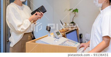 A scene of a woman wearing a mask holding a wallet and checking out at a reception desk with an acrylic board installed. A scene of a woman wearing a mask holding a wallet and checking out at a reception desk with an acrylic board installed. 93136431