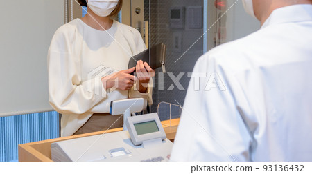 A scene of a woman wearing a mask holding a wallet and checking out at a reception desk with an acrylic board installed. A scene of a woman wearing a mask holding a wallet and checking out at a reception desk with an acrylic board installed. 93136432
