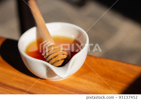Honey with dipper in small white bowl. Homemade honey with wooden stick in sunlight. Honey in jar. Natural sweetener. Healthy nutrition. Natural dessert concept. Sweet food. 93137452
