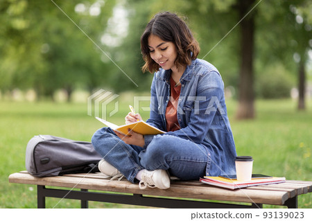 Arab female student writing in notebook while sitting on bench in park 93139023