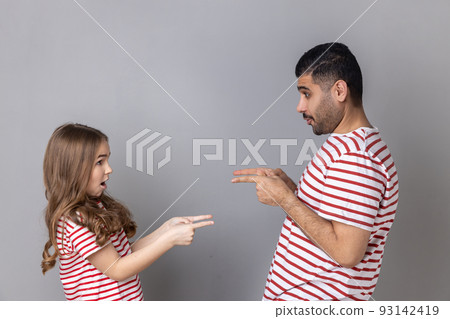 Portrait of surprised astonished father and daughter in striped T-shirts standing pointing at each other, looking with big eyes, choosing turn. Indoor studio shot isolated on gray background. Portrait of surprised astonished father and daughter in striped T-shirts standing pointing at each other, looking with big eyes, choosing turn. Indoor studio shot isolated on gray background. 93142419