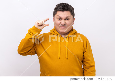 Portrait of funny man making little bit gesture with fingers and smiling sarcastic, showing minimum size, begging few more, wearing urban style hoodie. Indoor studio shot isolated on white background. 93142489