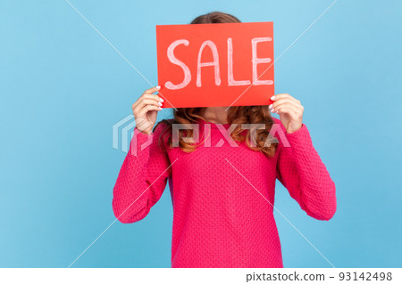 Portrait of unknown woman wearing pink pullover, covering face with Sale inscription, market discounts, black friday, low prices. Indoor her studio shot isolated on blue background. Portrait of unknown woman wearing pink pullover, covering face with Sale inscription, market discounts, black friday, low prices. Indoor her studio shot isolated on blue background. 93142498