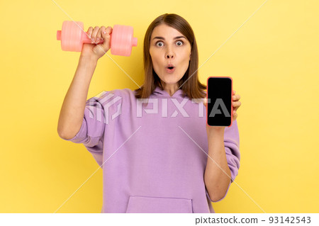 Shocked surprised woman holding blank screen smartphone and pink dumbbell, advertising new fitness application, wearing purple hoodie. Indoor studio shot isolated on yellow background. Shocked surprised woman holding blank screen smartphone and pink dumbbell, advertising new fitness application, wearing purple hoodie. Indoor studio shot isolated on yellow background. 93142543