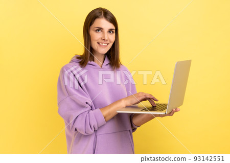 Portrait of optimistic positive young adult woman working on laptop compute, looking at camera with happy face, wearing purple hoodie. Indoor studio shot isolated on yellow background. 93142551