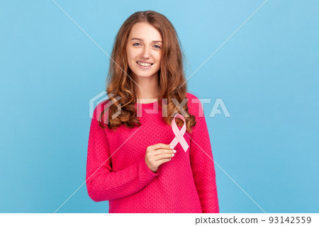 Lovely beautiful woman wearing pink pullover, holding pink ribbon, breast cancer awareness, female health diagnostics and cancer prevention. Indoor studio shot isolated on blue background. Lovely beautiful woman wearing pink pullover, holding pink ribbon, breast cancer awareness, female health diagnostics and cancer prevention. Indoor studio shot isolated on blue background. 93142559