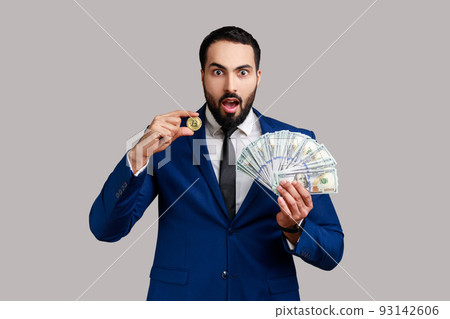 Shocked man showing dollar bills and golden coin of crypto currency, transferring money into digital bitcoins, wearing official style suit. Indoor studio shot isolated on gray background. Shocked man showing dollar bills and golden coin of crypto currency, transferring money into digital bitcoins, wearing official style suit. Indoor studio shot isolated on gray background. 93142606