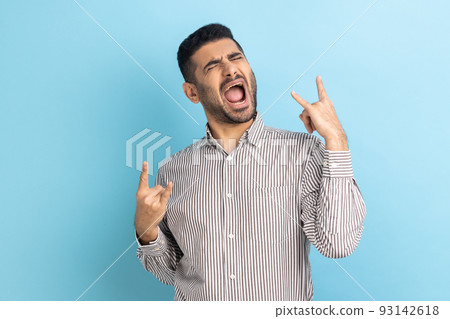 Disobedient businessman with beard showing rock and roll gesture and excitedly screaming, having fun, enjoying heavy music, wearing striped shirt. Indoor studio shot isolated on blue background. 93142618