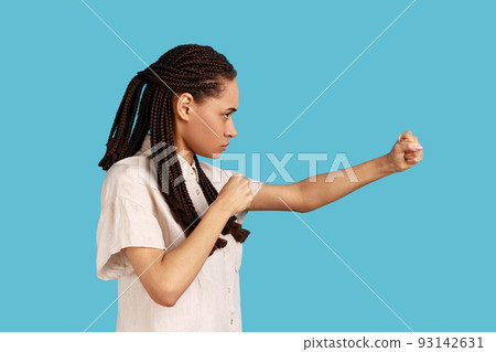 Side view portrait of woman with black dreadlocks having serious look, keeping fists clenched, defencing herself from offence and bad treatment. Indoor studio shot isolated on blue background. 93142631