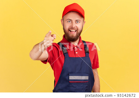Portrait of smiling bearded satisfied man worker standing and pointing finger to camera, choosing you, wearing overalls and red cap. Indoor studio shot isolated on yellow background. Portrait of smiling bearded satisfied man worker standing and pointing finger to camera, choosing you, wearing overalls and red cap. Indoor studio shot isolated on yellow background. 93142672