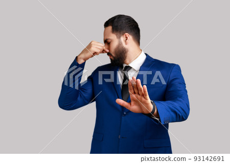 Bearded man grabbing nose, grimacing in disgust and showing stop gesture, expressing repulsion to stink, fart gases, wearing official style suit. Indoor studio shot isolated on gray background. Bearded man grabbing nose, grimacing in disgust and showing stop gesture, expressing repulsion to stink, fart gases, wearing official style suit. Indoor studio shot isolated on gray background. 93142691