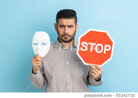 Portrait of serious businessman holding white mask with unknown face and red traffic sign, looking at camera, wearing striped shirt. Indoor studio shot isolated on blue background. Portrait of serious businessman holding white mask with unknown face and red traffic sign, looking at camera, wearing striped shirt. Indoor studio shot isolated on blue background. 93142710