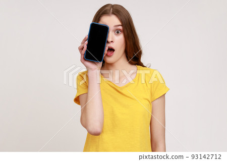 Portrait of excited shocked teenager girl wearing yellow T-shirt covering half face with cell phone and looking at camera with open mouth and big eyes. Indoor studio shot isolated on gray background. Portrait of excited shocked teenager girl wearing yellow T-shirt covering half face with cell phone and looking at camera with open mouth and big eyes. Indoor studio shot isolated on gray background. 93142712