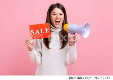 Excited brunette female holding sale card and megaphone, announcing discounts in shopping mall, wearing white casual style sweater. Indoor studio shot isolated on pink background. Excited brunette female holding sale card and megaphone, announcing discounts in shopping mall, wearing white casual style sweater. Indoor studio shot isolated on pink background. 93142715